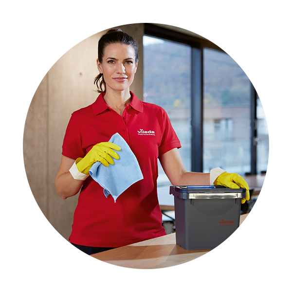 Une femme portant un polo rouge avec le logo "Vileda Professional", des gants de nettoyage jaunes et un chiffon en microfibre bleu. Elle se tient derrière une table en bois, son autre main reposant sur un seau de nettoyage Vileda gris. L'arrière-plan montre un intérieur moderne et lumineux avec de grandes fenêtres. L'image est recadrée dans un cadre circulaire sur un fond gris clair.