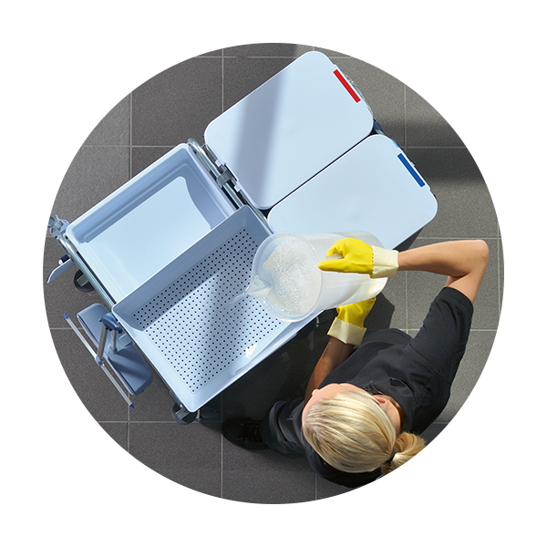Vue de haut d'une personne aux cheveux blonds, portant des gants jaunes et une chemise noire, versant un liquide savonneux d'un pichet transparent dans un chariot de nettoyage blanc spécialisé. Le chariot comporte plusieurs compartiments, dont un plateau perforé et deux bacs dont les couvercles sont codés en rouge et en bleu.