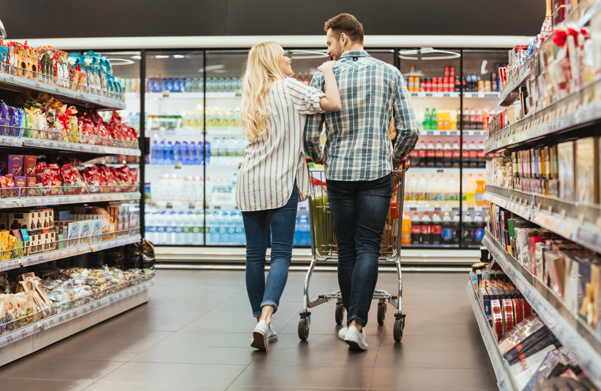 A couple, seen from behind, walks down a supermarket aisle with a shopping cart. The woman has long blonde hair and is wearing a striped shirt and jeans, resting her hand on the man's shoulder as they look at each other. The man is wearing a plaid shirt and jeans. The aisle is lined with shelves of snacks and refrigerated glass cases filled with bottled drinks.